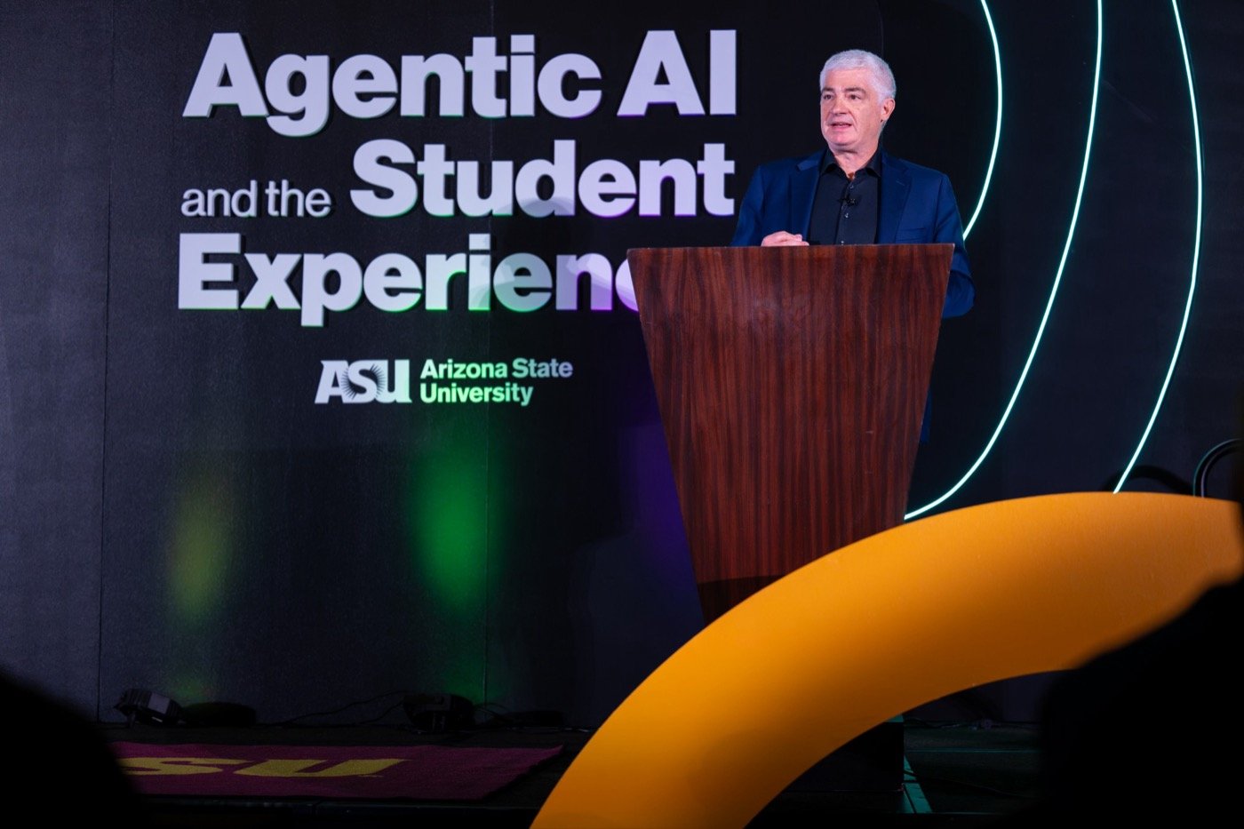 A speaker at the podium on the Agentic AI and the Student Experience main stage, with the event title on the backdrop, glowing neon arcs, and a curved gold set piece in front