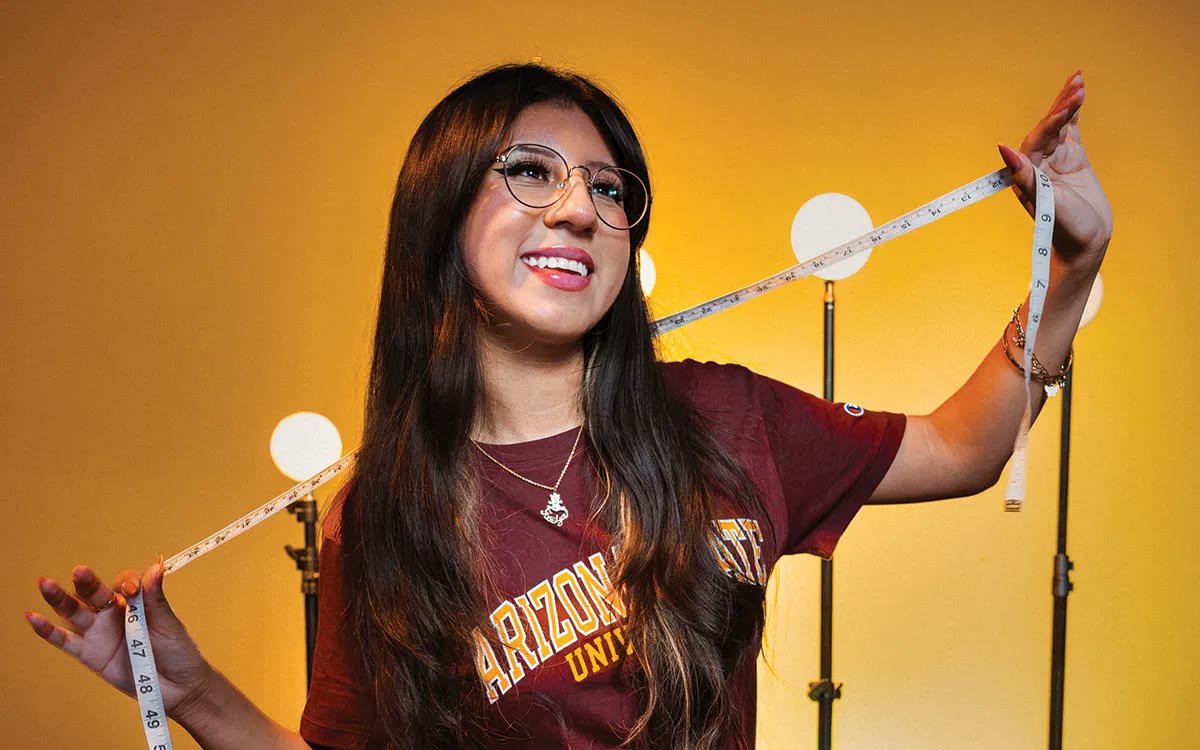 An ASU student in a maroon Arizona State tee smiles while stretching a tape measure between her hands, lit gold against studio lights, from the Volume 5 cover shoot