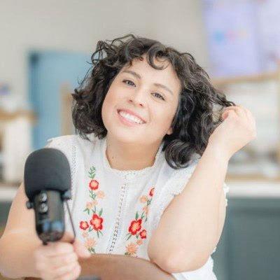 Alisha Mendez smiling with her head tilted, leaning toward a podcast microphone, in a white top with red embroidered flowers