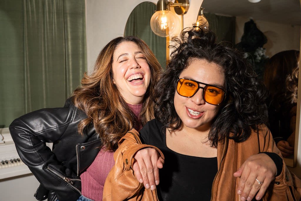 The hosts laughing together, one in orange sunglasses and a tan leather jacket, in a room with green curtains and a piano