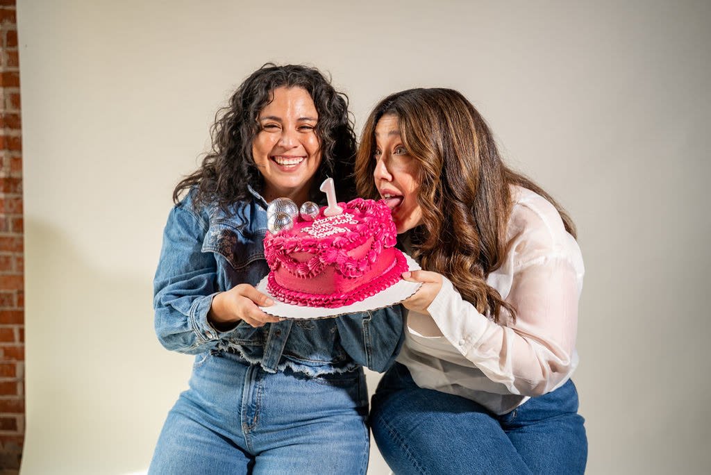 The hosts holding a pink heart-shaped birthday cake with a number one candle and disco ball toppers