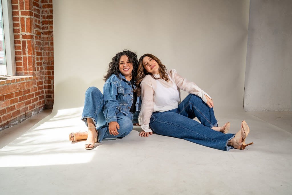 The hosts sitting on the floor of a sunlit studio with brick walls, leaning into each other