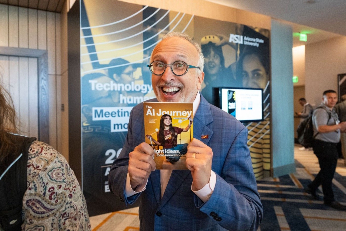 Lev Gonick grinning while holding up The AI Journey Volume 5 in front of the entry wall