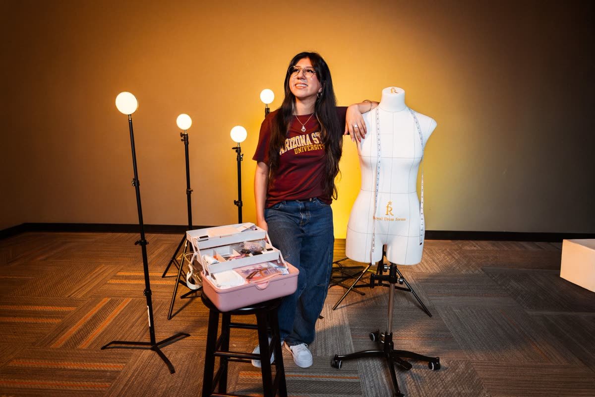 The cover subject leans on a dress form beside a sewing kit on a stool, surrounded by globe lights