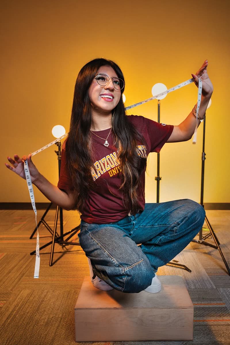 The cover subject crouches on a wooden box, stretching a tape measure between her hands in front of glowing globe lights